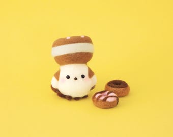 May include: A needle-felted bird figurine with a brown and white cap resembling a mushroom, featuring black bead eyes and pink cheeks, accompanied by two small felted donut shapes in brown and pink on a solid yellow background.