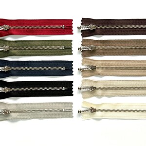 Op de afbeelding: Assortiment van 10 metalen ritsen in verschillende kleuren, waaronder rood, bruin, groen, beige, zwart en wit. Elke rits heeft een zilveren schuif en is ongeveer 15 cm lang.