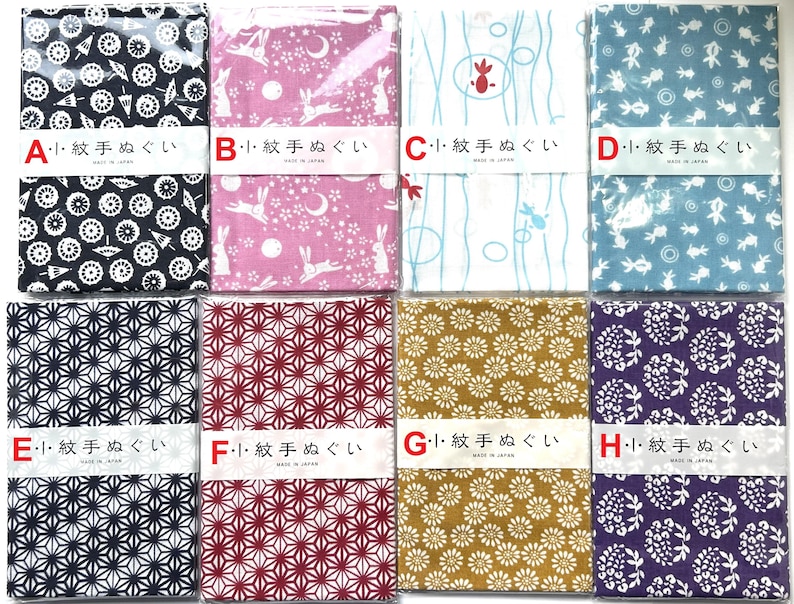May include: Eight rectangular tenugui cloths arranged in two rows, each wrapped in plastic with a paper band labeled A through H. The patterns include white motifs on black, pink, blue, and purple backgrounds, featuring umbrellas, rabbits, fish, and flowers.