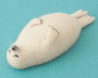 May include: A handmade, off-white felt seal figurine lying on its back. The seal has black dots for eyes and flipper details, with a small black nose and white whiskers. The background is a solid light blue.