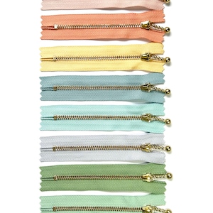 Könnte beinhalten: Ein Set mit 12 Reißverschlussbändern in verschiedenen Pastellfarben, darunter Rosa, Pfirsich, Gelb, Blau, Grün und Grau. Jedes Reißverschlussband hat einen goldenen Metallreißverschluss mit einer kleinen Kugel am Ende.