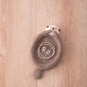 Puede incluir: Una nutria de fieltro marrón con la cara blanca y los ojos negros, enroscada en forma de cuenco. La nutria sostiene una pequeña cadena de plata con dos abalorios azules y blancos en su interior.