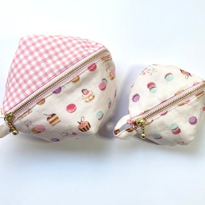 May include: Two pink and white fabric zippered pouches with a gingham pattern on one side and a pastel macaron and cake pattern on the other side.
