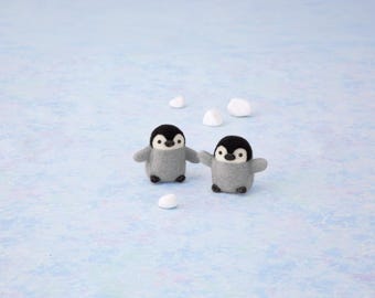 May include: Two small needle-felted penguin figurines with black heads, white faces, and grey bodies. The penguins have small black eyes and beaks, and are positioned near several small, smooth white stones on a light blue surface.