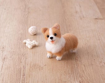 May include: A handcrafted felt corgi dog figurine with a white and tan coat, black eyes, and a pink tongue. Beside the dog are two small, white, textured objects. The figurine is set on a light brown wooden surface.