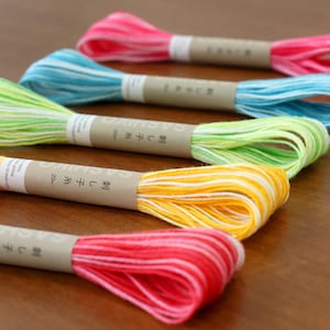 May include: Close-up of five skeins of colorful embroidery floss. The floss is in shades of pink, blue, green, yellow, and red. Each skein is wrapped with a paper band that has Japanese text. The floss is 20m in length.