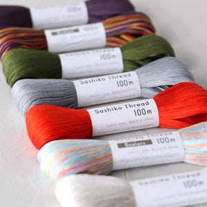 Peut inclure: Assortiment de bobines de fil Sashiko de différentes couleurs, dont rouge, orange, gris, vert et multicolore. Chaque bobine est étiquetée "Sashiko Thread" et indique la longueur du fil en mètres. L'adresse du site Web "https://olympus-thread.com" est imprimée sur chaque étiquette.