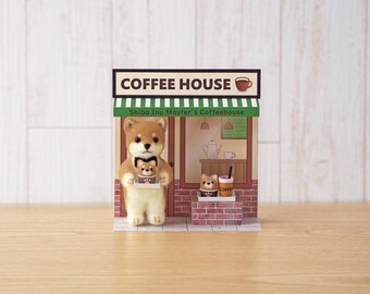 May include: A miniature diorama of a coffee house featuring a felt Shiba Inu figure holding a tray with a smaller dog figurine. The shop front has a green and white striped awning, a sign reading COFFEE HOUSE, and Shiba Inu Master's Coffeehouse text. A brick base displays another small dog figure and a coffee cup.