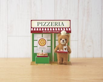 May include: A felt bear figurine wearing a white chef hat stands in front of a miniature pizzeria storefront. The bear holds a tray with a drink and pizza. The shop features green window frames, a red and white striped awning, and signs reading PIZZERIA and BEAR CHEF'S PIZZERIA.