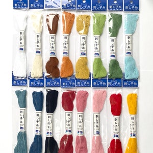 May include: Assortment of 18 skeins of embroidery floss in various colours, including white, beige, brown, orange, yellow, green, blue, red, pink, and yellow. Each skein is labelled "オリムパス 刺し子糸" which translates to "Olympus Sashiko Thread".