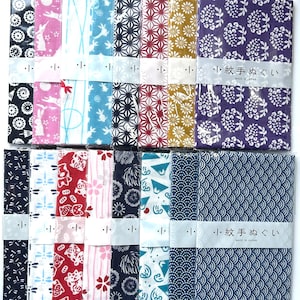 May include: A collection of sixteen rectangular cotton cloths, known as tenugui, arranged in two rows. Each cloth features distinct patterns including rabbits, flowers, geometric shapes, and waves, all wrapped in clear plastic with a paper band.