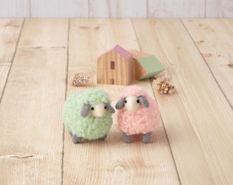 May include: Two needle-felted sheep figurines, one in light green and one in light pink, each featuring grey ears, grey legs, and a cream-colored face with black eyes. Behind the sheep are two small wooden house-shaped blocks.