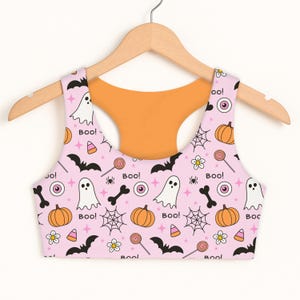May include: A light pink racerback sports bra featuring a repeating Halloween pattern of white ghosts, orange pumpkins, black bats, spiderwebs, candy corn, lollipops, bones, eyeballs, and flowers with the text Boo! printed in black throughout the fabric.