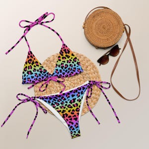 May include: A two-piece bikini set with a vibrant rainbow leopard print. The top is a triangle style with tie straps, and the bottoms are a classic cut with tie sides. A round woven bag, sunglasses, and a woven mat are also in the image.