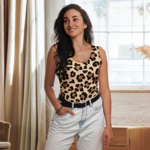 May include: A sleeveless bodysuit with a tan background and a brown leopard print. The bodysuit is paired with light-wash jeans and a black belt. The model is wearing a necklace and bracelet.