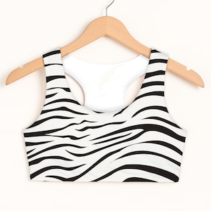 May include: A white and black zebra print sports bra with a racerback design. The bra is hanging on a wooden hanger against a white background. The zebra stripes are black and wavy.