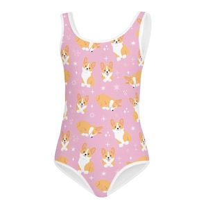 May include: A pink one-piece swimsuit featuring a repeating pattern of cartoon corgi dogs in various poses. The dogs are primarily orange and white, set against a pink background with white accents. The swimsuit has white trim.