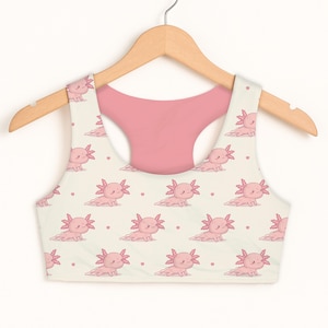 May include: A cream-colored sports bra with a pink axolotl pattern. The axolotls are pink with detailed gills and are scattered across the front. The back of the bra is solid pink. The bra is hanging on a wooden hanger.