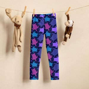 May include: Purple, blue and pink sea turtle leggings on a white background. The leggings are hanging on a clothesline with a stuffed animal on each side.