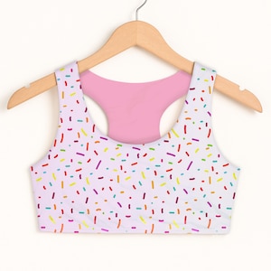 May include: A white sports bra with a colorful sprinkle pattern. The bra has a racerback design with a pink interior and is hanging on a wooden hanger.