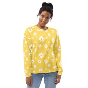 May include: Yellow sweatshirt with a daisy pattern. The sweatshirt features a repeating pattern of white daisies with yellow centers. The long-sleeved top is worn by a person wearing blue jeans.