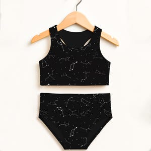 May include: Black two-piece swimsuit set with a constellation pattern. The top is a racerback style, and the bottoms are high-waisted. The white constellation design is set against a black background. The swimsuit is hanging on a wooden hanger.