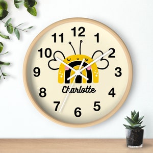 May include: A yellow and black bee with a rainbow on its back is featured on a cream-colored clock with black numbers. The clock has a wooden frame and the name "Charlotte" is printed below the bee.