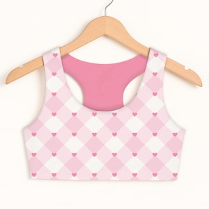 May include: A pink and white sports bra with a heart pattern. The bra is hanging on a wooden hanger. The pattern consists of pink hearts on a white and pink checkered background. The back of the bra is solid pink.