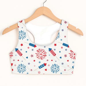 May include: White sports bra with a patriotic pattern of red and blue fireworks, stars, and hearts. The bra is hanging on a wooden hanger. The design is repeated across the fabric.