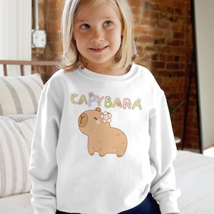 May include: White sweatshirt featuring a cartoon capybara with a flower and the word "CAPYBARA" in colorful letters. The capybara is light brown with a flower on its head. The sweatshirt is a crew neck style.
