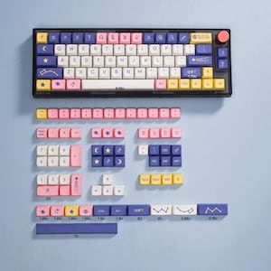 May include: A keyboard with a blue, pink, and yellow color scheme. The keys have various symbols and designs, including stars, moons, and rockets. The keyboard is on a light blue background.