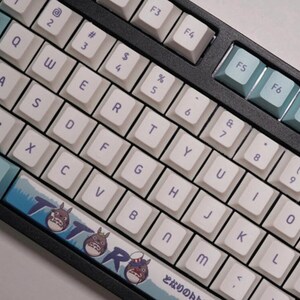 May include: A white mechanical keyboard with a black frame and blue and white keycaps. The keyboard has a custom keycap set with a Totoro design. The keyboard has a Japanese character on the bottom right keycap.