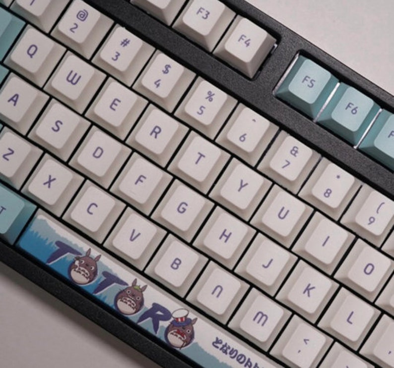 May include: A white mechanical keyboard with a black frame and blue and white keycaps. The keyboard has a custom keycap set with a Totoro design. The keyboard has a Japanese character on the bottom right keycap.