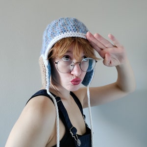 May include: A person wearing a blue and brown crocheted hat with white trim and white strings. The hat has ear flaps. The person is wearing round glasses and has red hair. They are saluting with their right hand.