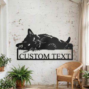 May include: A black metal wall sign featuring a silhouette of a cat lying down with its head resting on a rectangular frame. The frame contains the words CUSTOM TEXT in a bold, sans-serif font. The piece is designed for wall mounting.