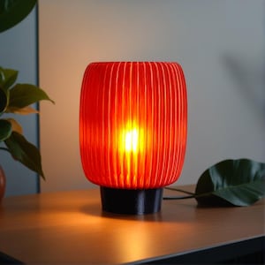 Peut inclure: Une lampe de table rouge avec un abat-jour à nervures et une base noire. La lampe est allumée et la lumière brille à travers l'abat-jour.