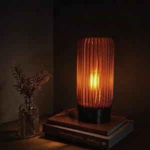 Peut inclure: Une lampe de table cylindrique avec un abat-jour en verre ambré strié et une base noire, diffusant une lueur chaude. La lampe est posée sur une pile de deux livres, dont l'un porte le texte 'baking with passion' sur la tranche, près d'un vase.