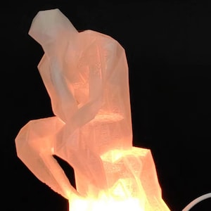 Puede incluir: Una escultura blanca translúcida impresa en 3D de una persona sentada con la cabeza en la mano. La escultura está iluminada desde adentro, proyectando un cálido resplandor naranja.