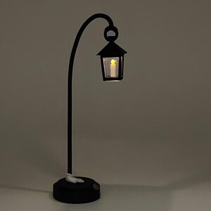 May include: A black metal table lamp with a curved arm and a small lantern-style shade. The lantern has a clear glass panel and a warm white LED light inside.
