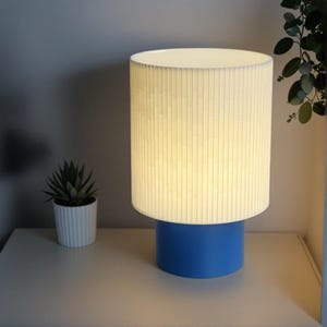 May include: A white lamp with a blue base. The lampshade has a ribbed texture and is lit up.