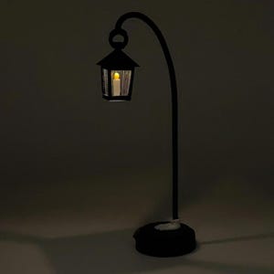 May include: A black metal lamp with a curved arm and a lantern-style shade. The lantern has a clear glass panel and a warm white light inside.