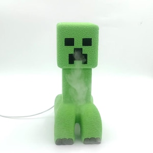 May include: A green, Minecraft-themed humidifier shaped like a Creeper. The humidifier has a textured surface and black square eyes and mouth. White vapor is emitted from the mouth. The humidifier is on a white background.