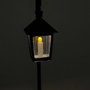 May include: A black metal street lamp with a clear glass lantern. A small, yellow LED light is lit inside the lantern, resembling a candle.