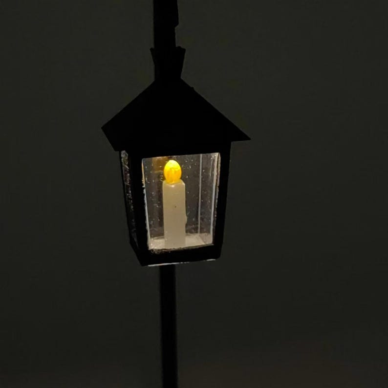 May include: A black metal street lamp with a clear glass lantern. A small, yellow LED light is lit inside the lantern, resembling a candle.