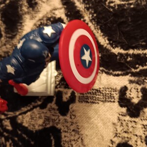 May include: A red, white, and blue plastic action figure of Captain America. The figure is holding a red, white, and blue shield.