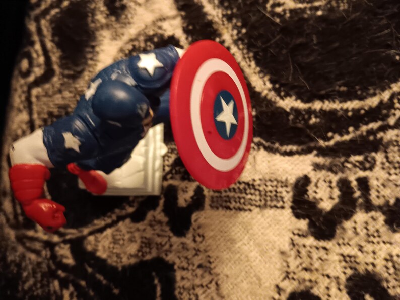May include: A red, white, and blue plastic action figure of Captain America. The figure is holding a red, white, and blue shield.
