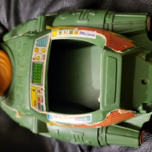 Puede incluir: Una nave espacial de juguete verde y naranja vintage, con una cabina y pegatinas que representan un panel de control. El juguete muestra signos de desgaste, con daños en los bordes. El diseño sugiere un tema de ciencia ficción retro.