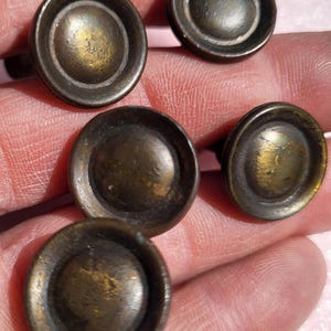 May include: A collection of five antique bronze-colored cabinet knobs. Each knob has a circular design with a raised center and a slightly recessed outer ring. The knobs show signs of age and wear, with variations in color and texture.