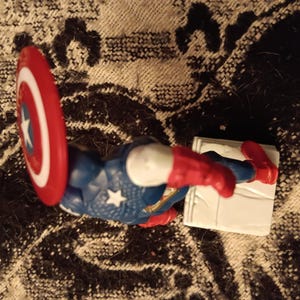 May include: A red, white, and blue action figure of Captain America with a red, white, and blue shield. The figure is standing on a white base.