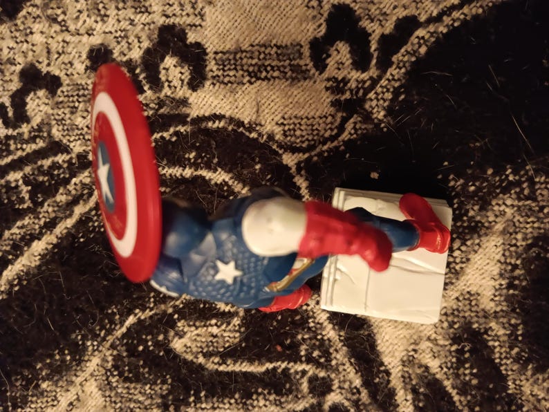 May include: A red, white, and blue action figure of Captain America with a red, white, and blue shield. The figure is standing on a white base.